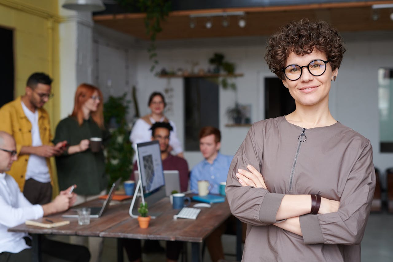 gallery-04 Smiling businesswoman with curly hair stands confidently in a modern office space with colleagues.