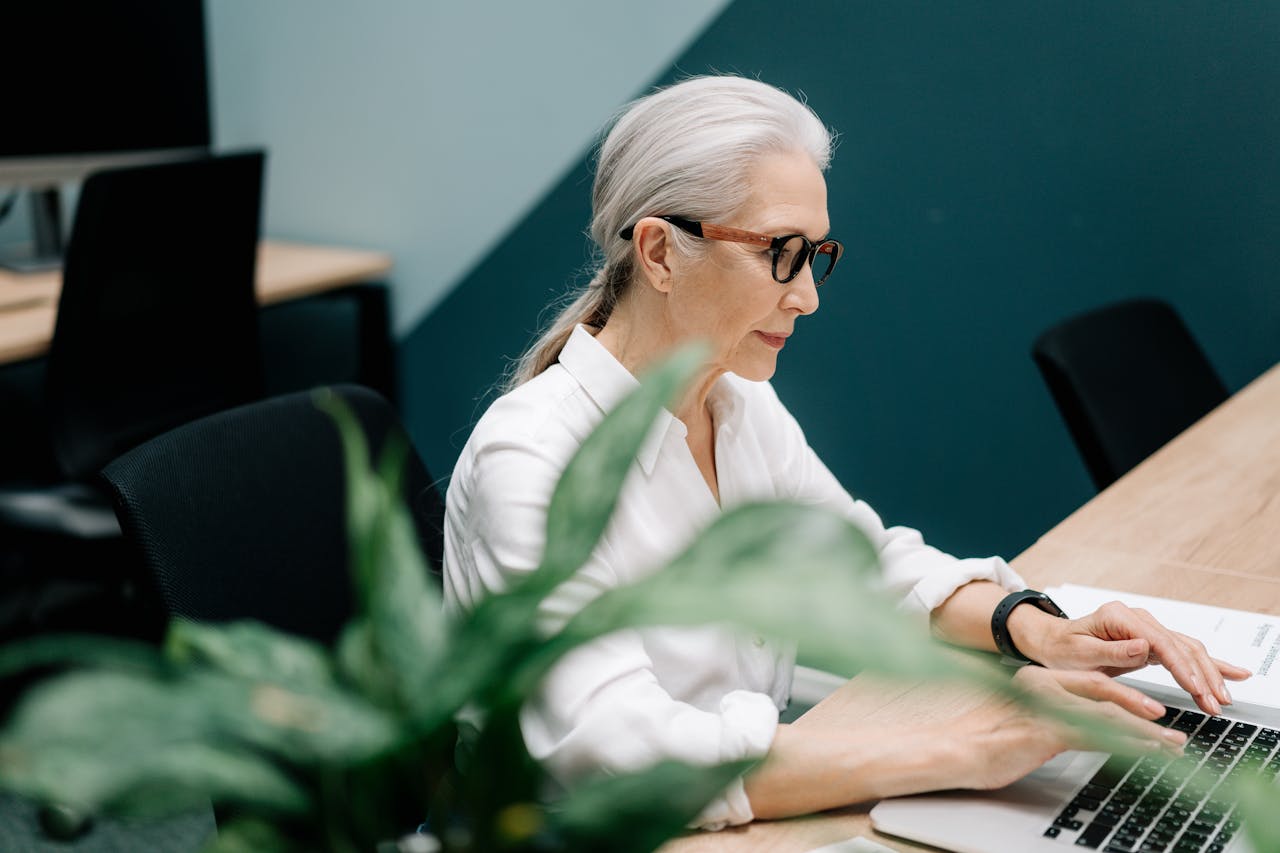 gallery-02 Confident senior woman with glasses working on a laptop in a modern office setting.